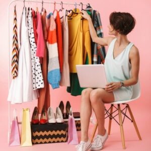 Photo of a woman sitting in front of a clothing wrack with a laptop on her knees. Make money online as a virtual personal stylist.