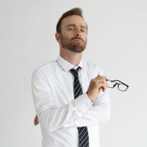 Photo of a man in a white dress shirt and tie, looking smug, arms crossed, holding glasses in one hand.