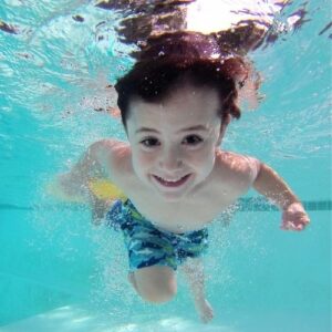 Close up photo of a smiling boy swimming underwater in a pool.