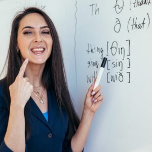 Close up photo of woman standing beside a whiteboard with words written on it, pointing to her open mouth.